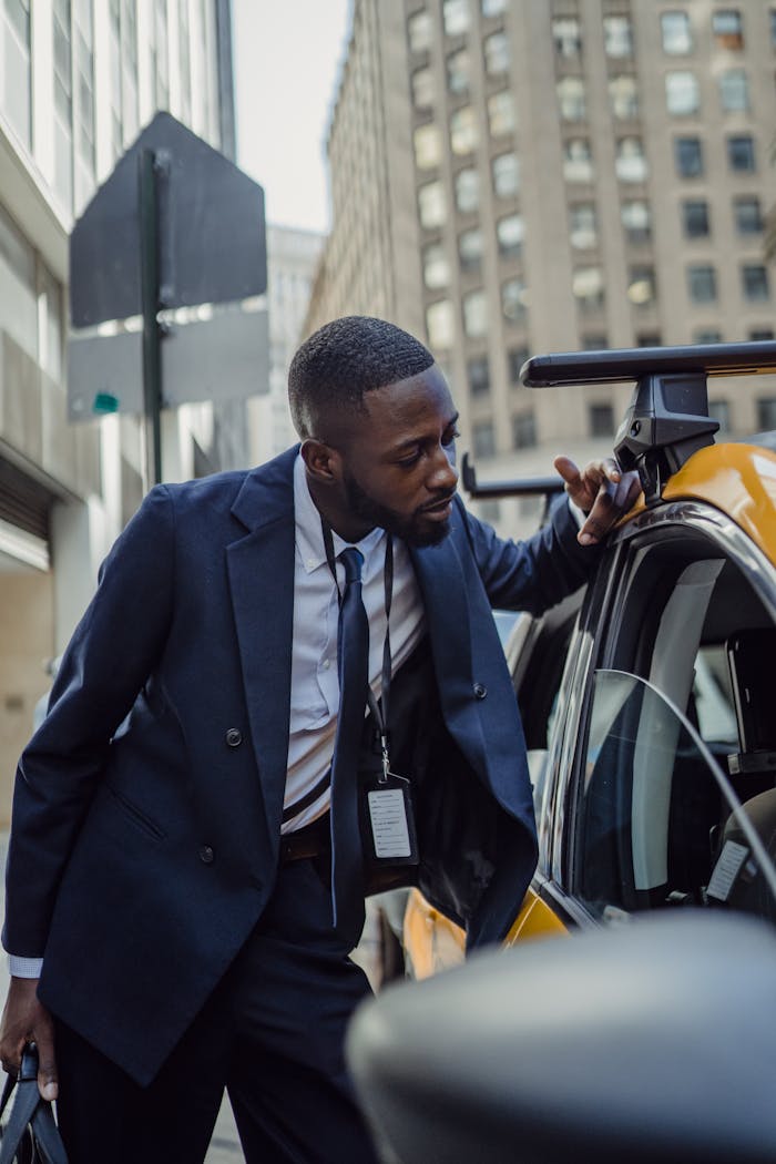 Man in formal attire leaning on a yellow taxi in an urban city environment.